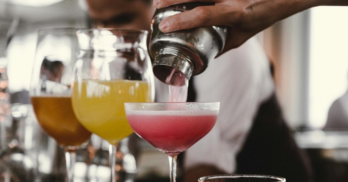 Three mixed drinks on a counter, with a shaker pouring a pink liquid into the far right drink.