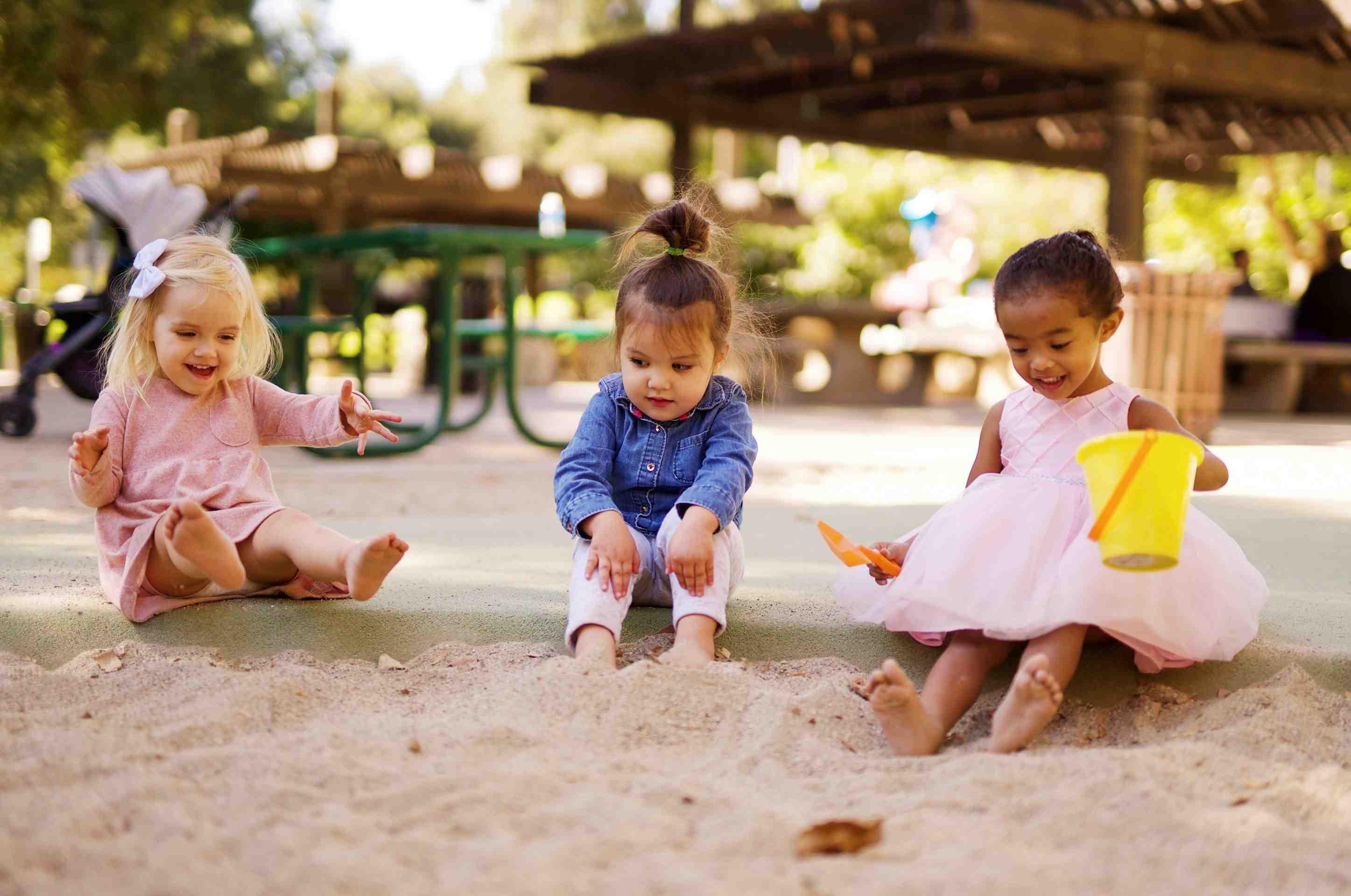 Three toddlers play together in a sandbox at a park.