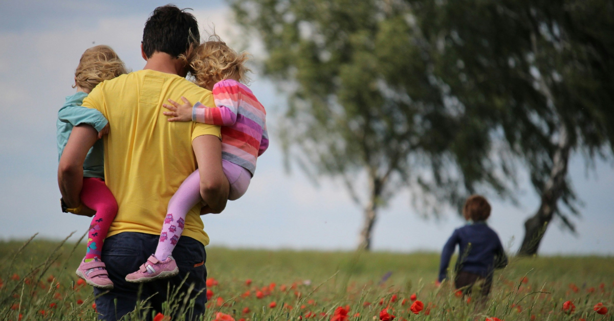A man carries two small girls in his arms in a field 