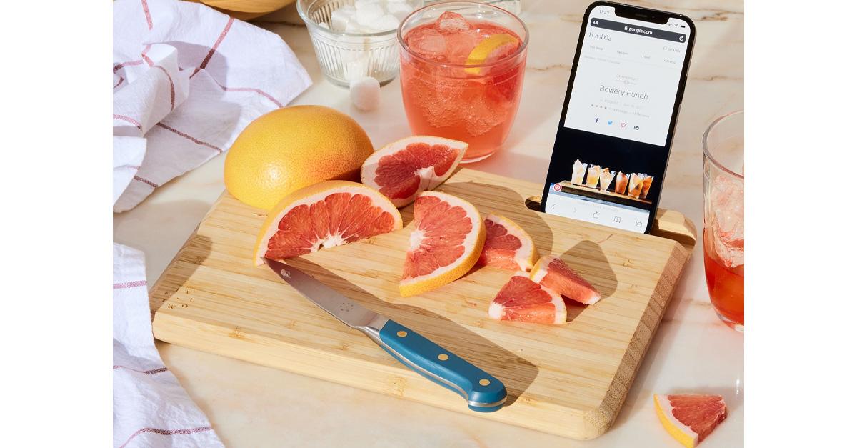 Bamboo cutting board with knife, a cut-up grapefruit, grapefruit cocktails and a smartphone