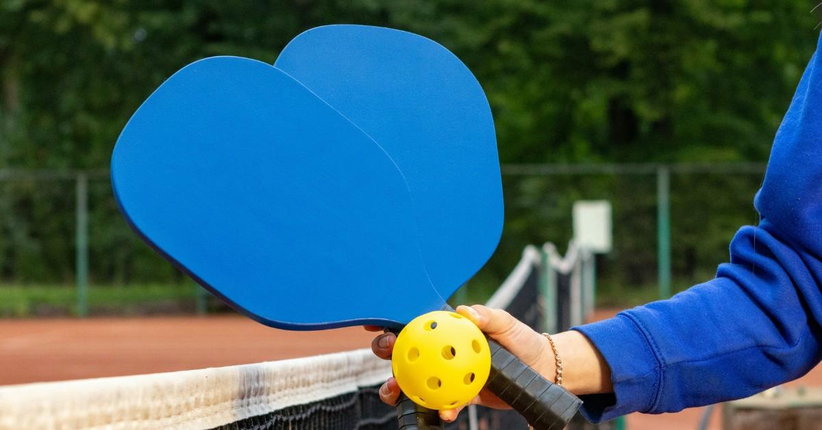 A person holds two blue pickleball paddles and a yellow ball