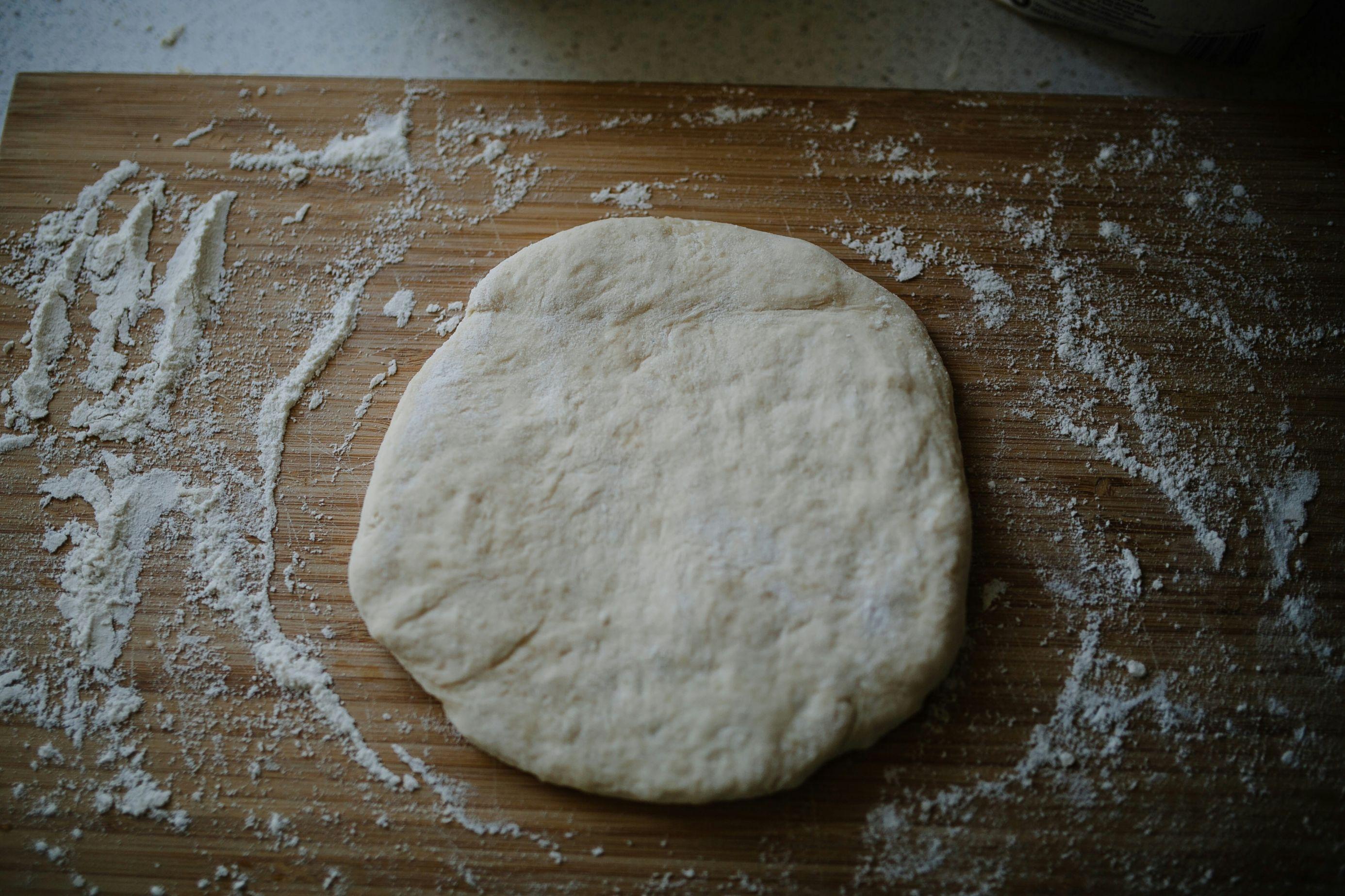 A picture of circular pizza dough atop a wooden table with flour all around the dough.