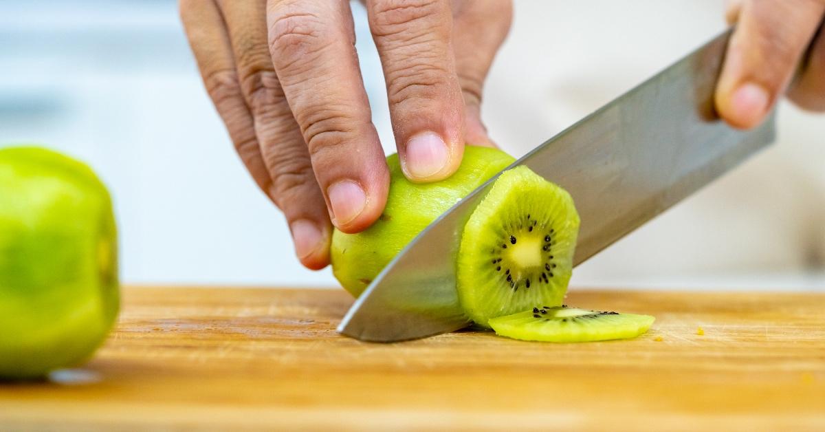 Person slicing Kiwi with a knife on a cutting board. 