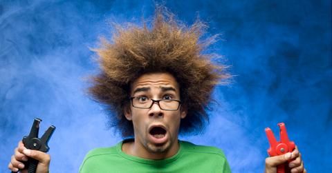 A man with an open mouth and shocked expression holds two electric plugs with his messy hair spiked every which way.