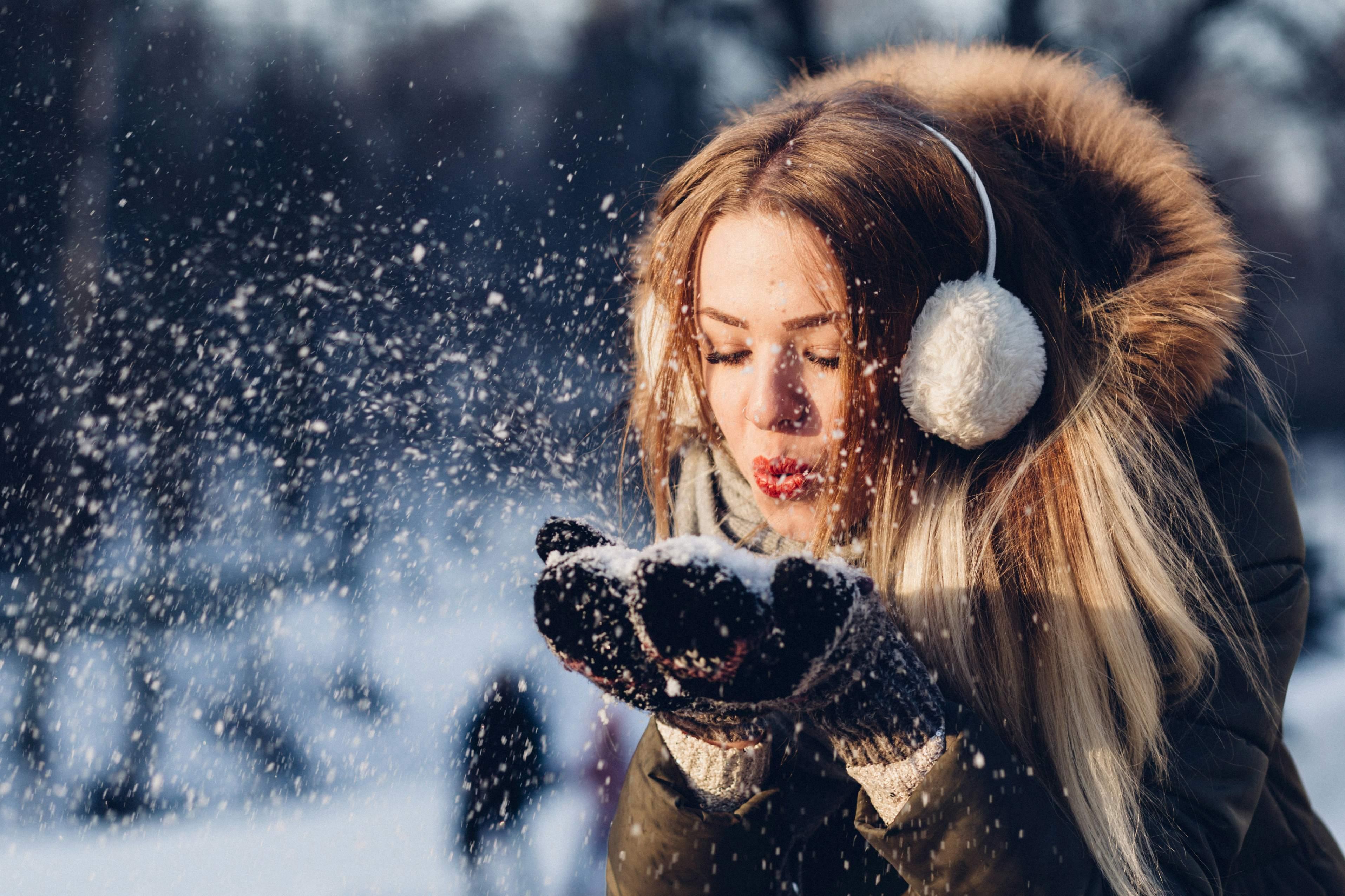 A woman wearing earmuffs and gloves blows snow from a shrinking snowball in her palms.