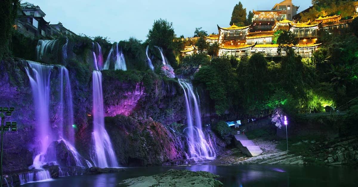 Furong Ancient Town's waterfall at night lit up with purple neon lighting and town lights around it.