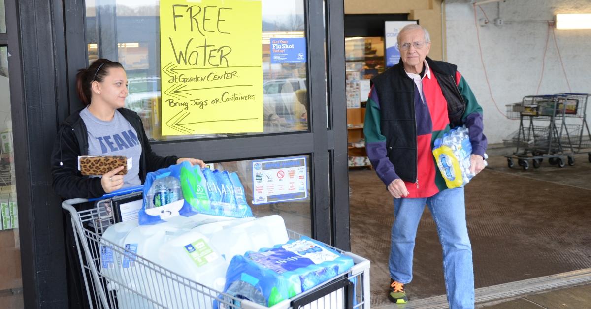 A woman stands in front of a sign reading "free water" with a shopping cart full of water, and a man exits a store, carrying a case of water.