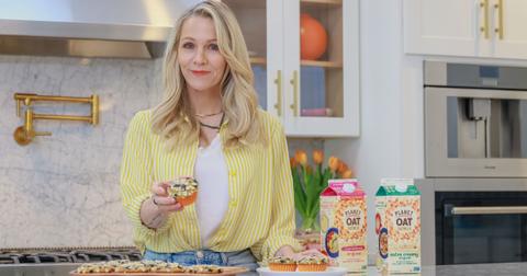 Photo of actor Jennie Garth holding a blueberry muffin beside containers of Planet Oat plant-based milk in a kitchen
