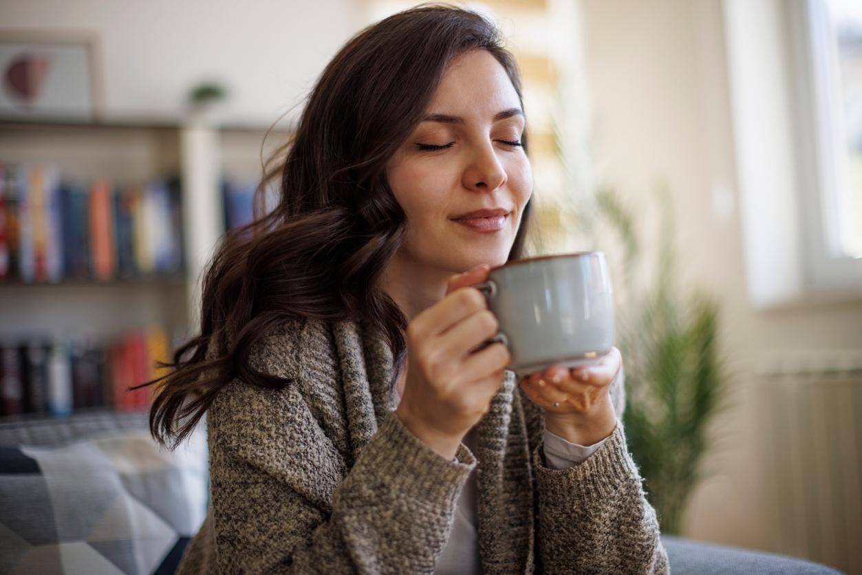 A woman in a sweater sips coffee from a cup in her living room.