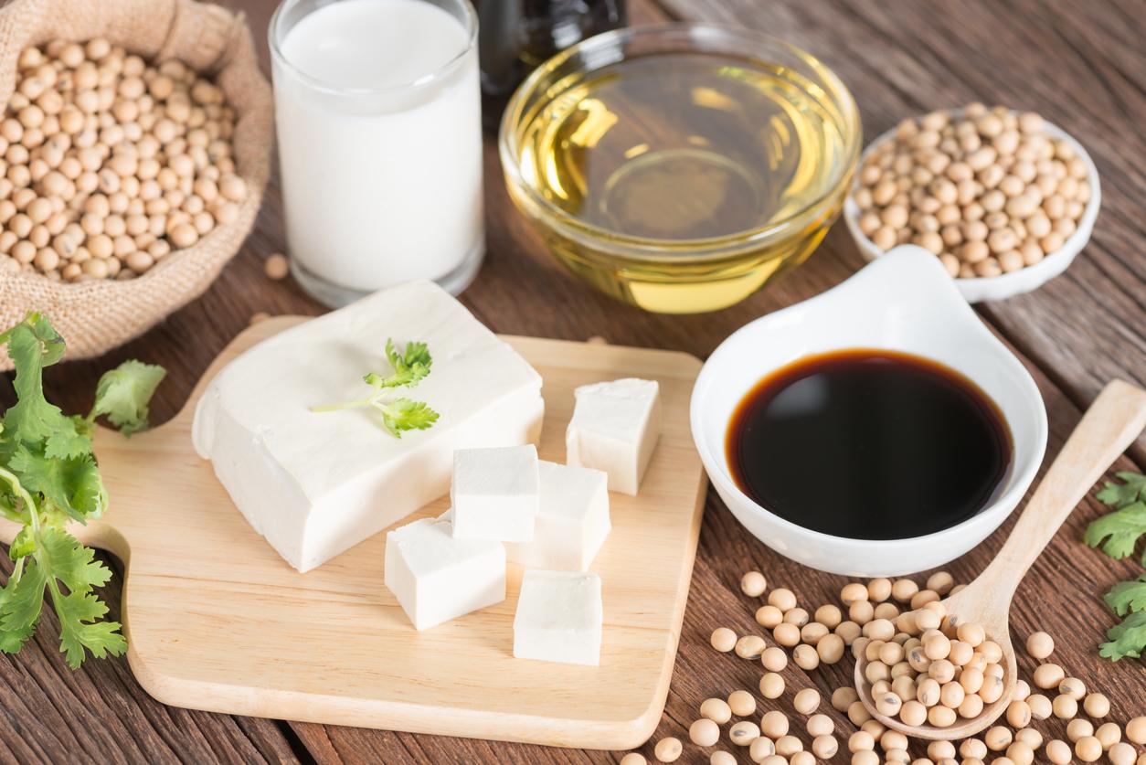 A block of tofu sits atop a cutting board beside soy sauce, soybeans, and oil.