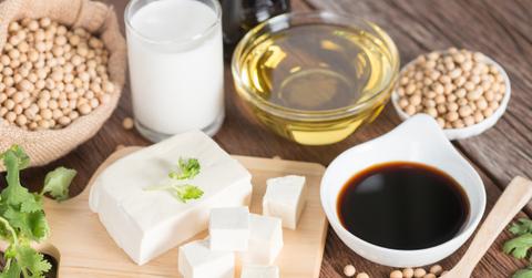 A block of tofu sits atop a cutting board beside soy sauce, soybeans, and oil.