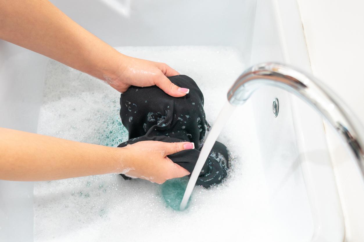 A side view of a person holding a wet, black clothing item in a tub of soapy water with the spout turned on above it.