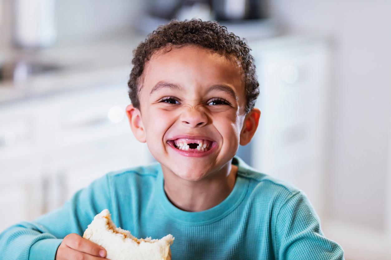 A smiling child in a blue shirt with teeth missing smiles while holding a peanut butter sandwich.