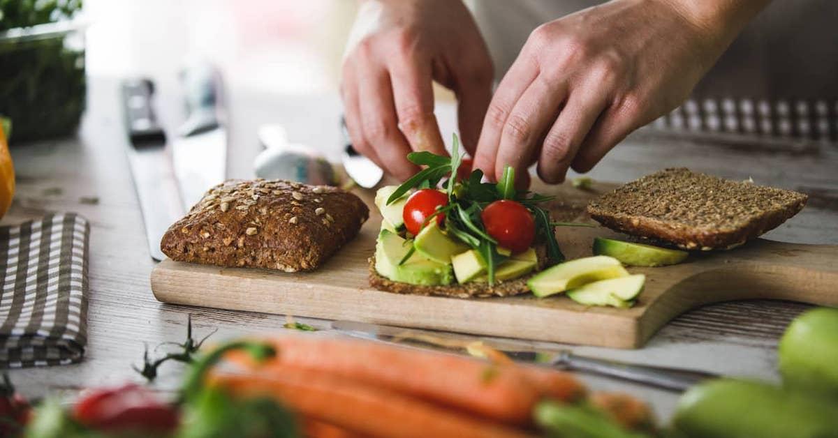 Closeup of a person preparing an avocado, tomato, and rocket sandwich on a bread board.