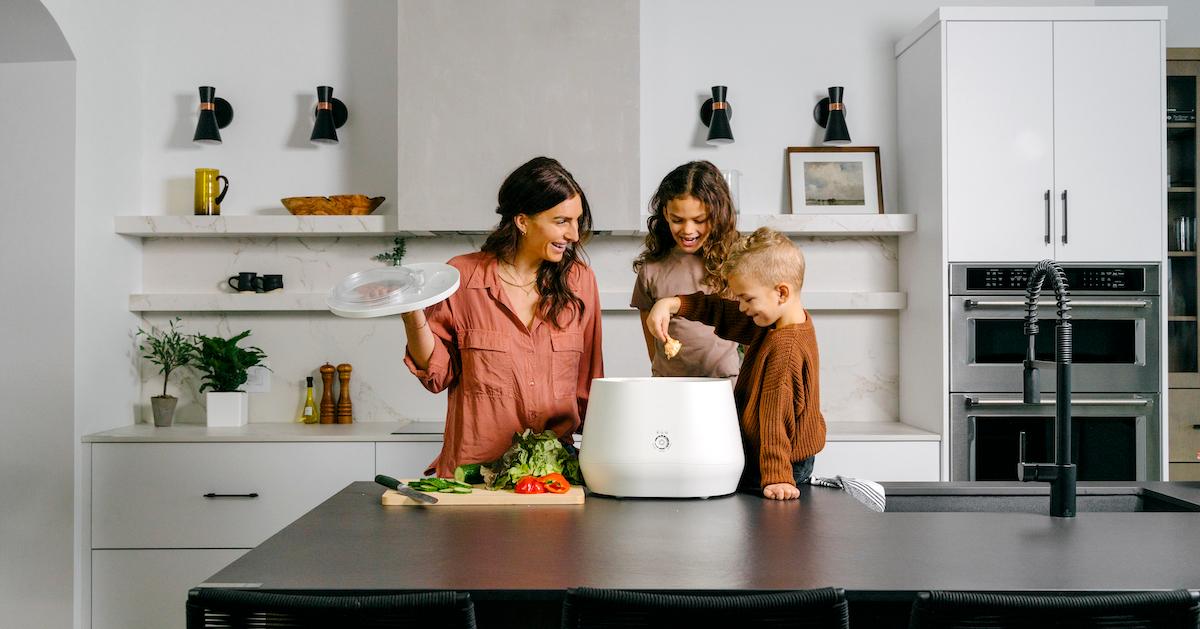 An adult and two young children stand at a kitchen counter and place a food scrap into a white Lomi composting machine.