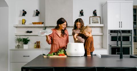 An adult and two young children stand at a kitchen counter and place a food scrap into a white Lomi composting machine.