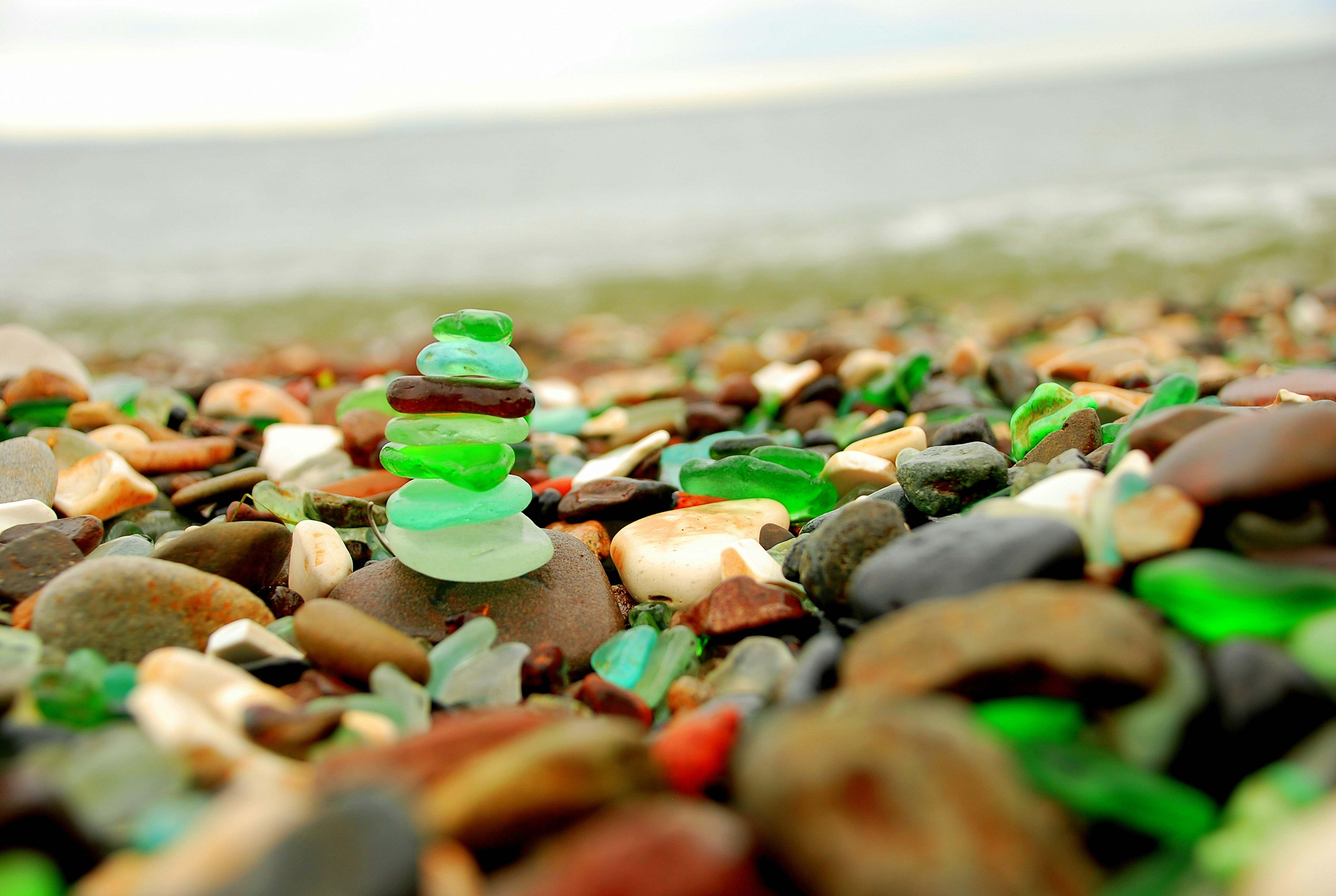 A collection of colorful sea glass is stacked atop one another amidst pebbles and rocks of various colors by a body of water.