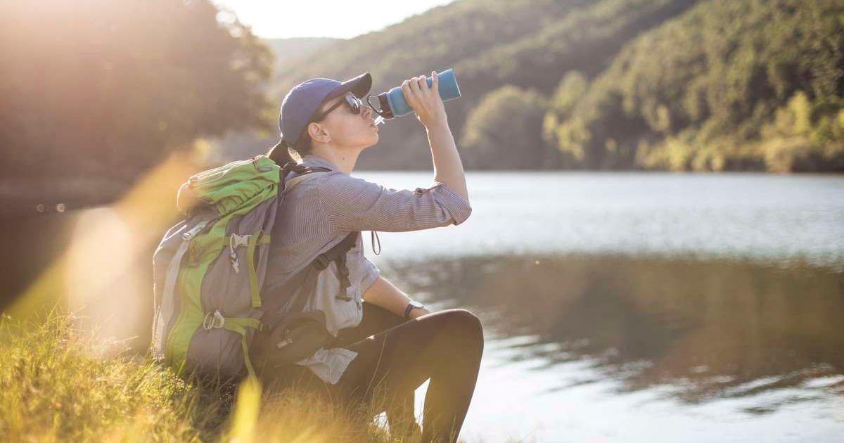 Woman drinking water from a bottle in front of a lake in Yellowstone National Park (Representative Cover Image Source: Getty Images | Vladimir Timotijevic)
