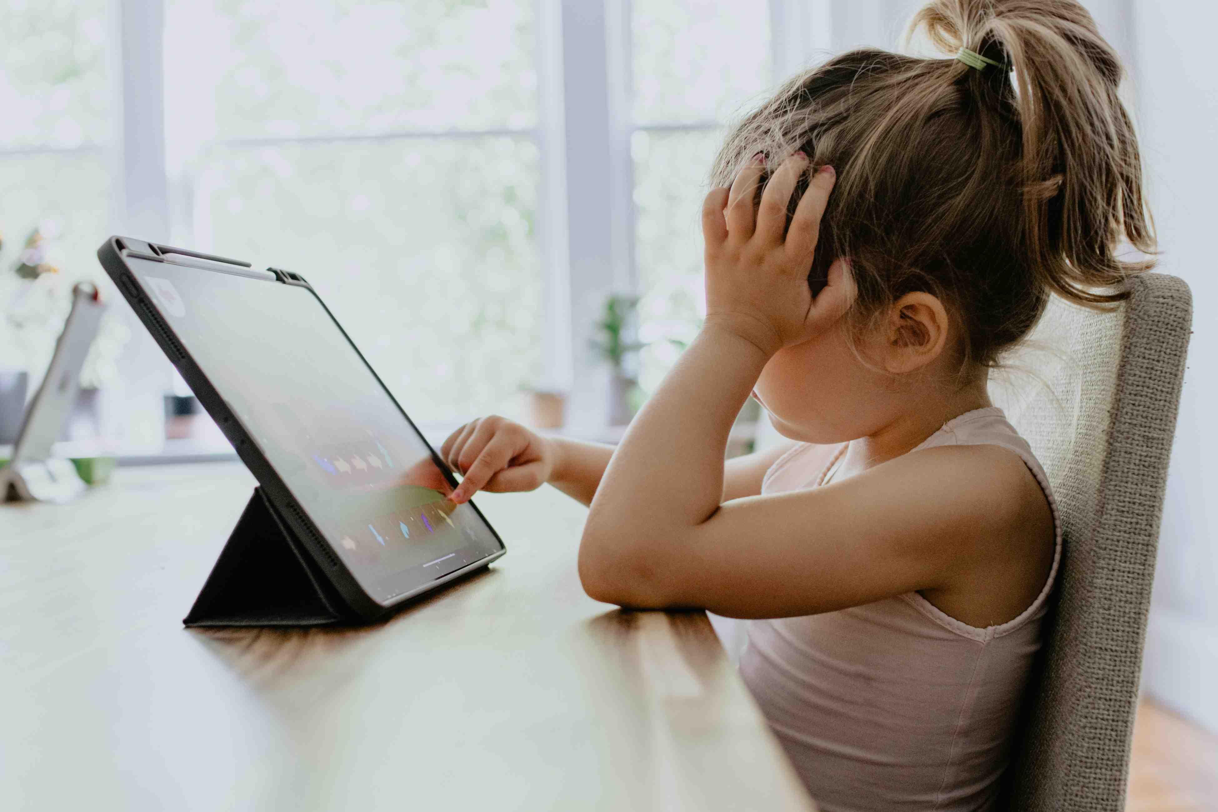 A young girl sitting in a chair at the dining room table performs a school lesson on a tablet.