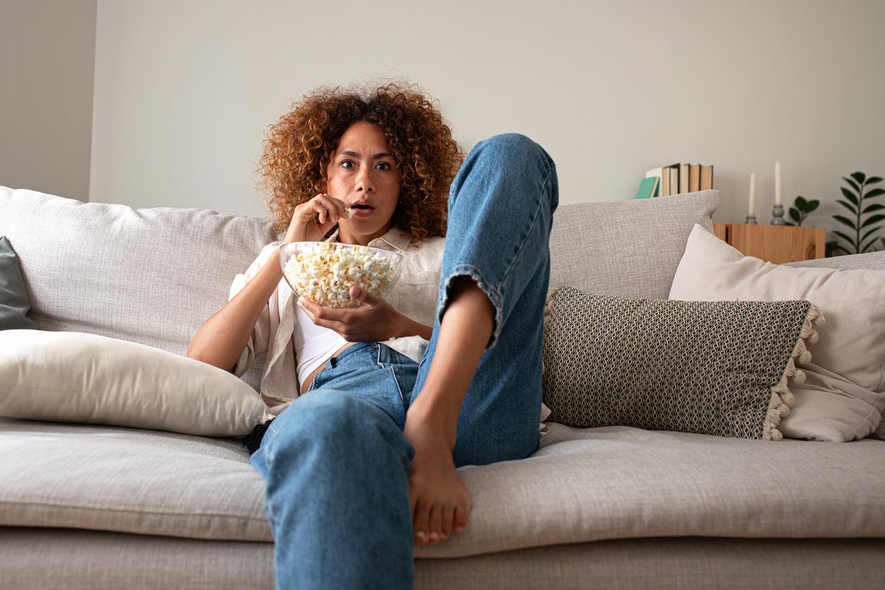 A Black woman sits on the couch watching television with a surprised look on her face