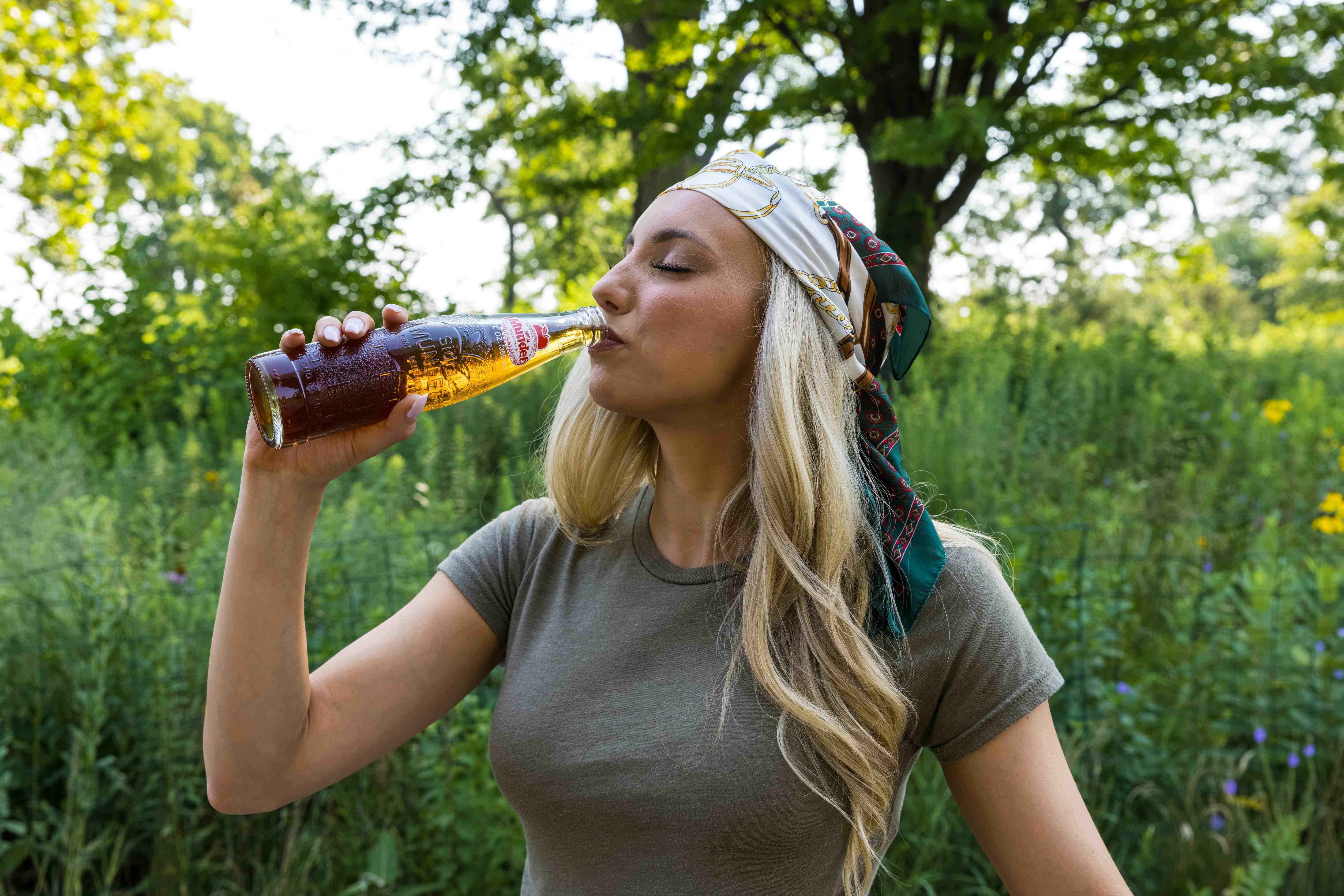 A woman takes a sip of a soft drink from a glass bottle while standing in nature.