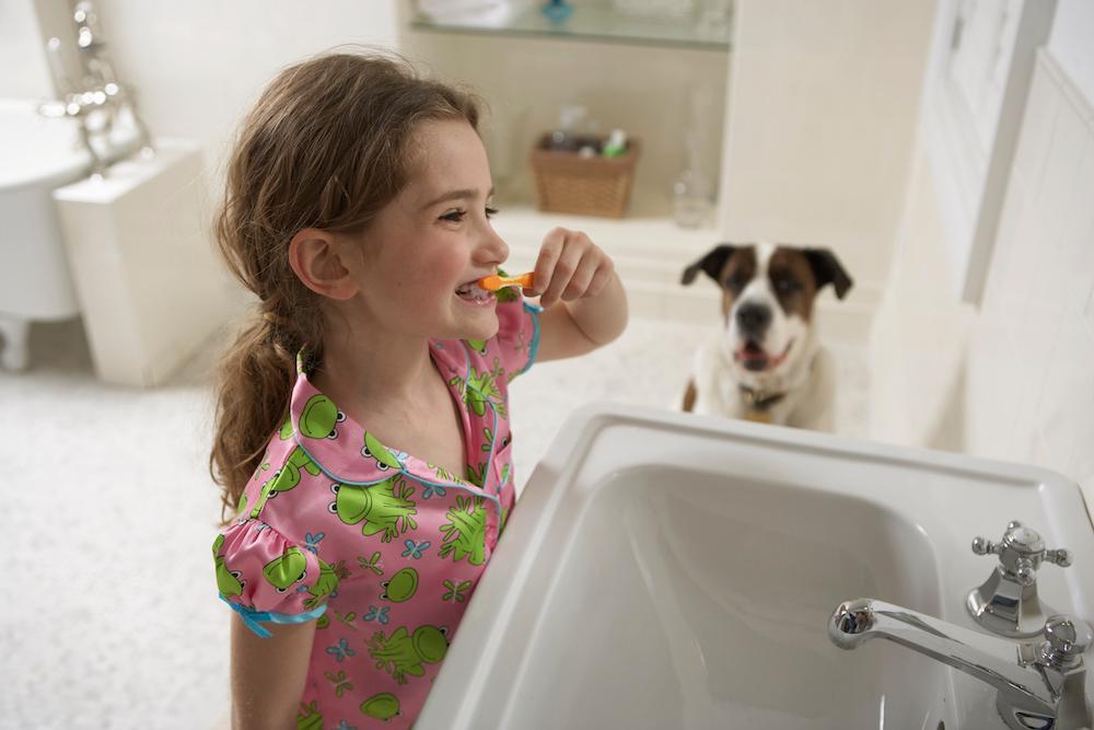 A young girl brushes her teeth while her dog watches. 