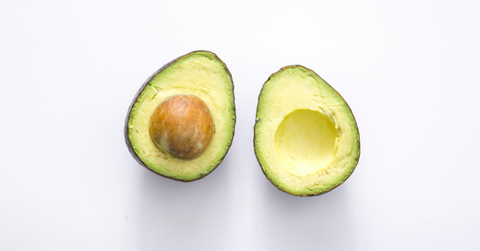 An avocado lays cut in half on a table with the pit remaining in one side