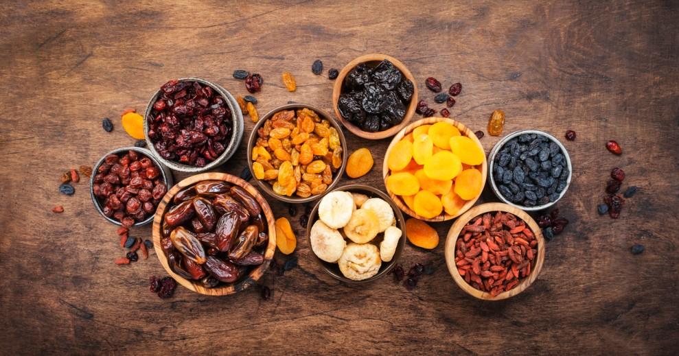 Nine bowls of varying sizes containing different dried fruits are pictured atop a wooden table.