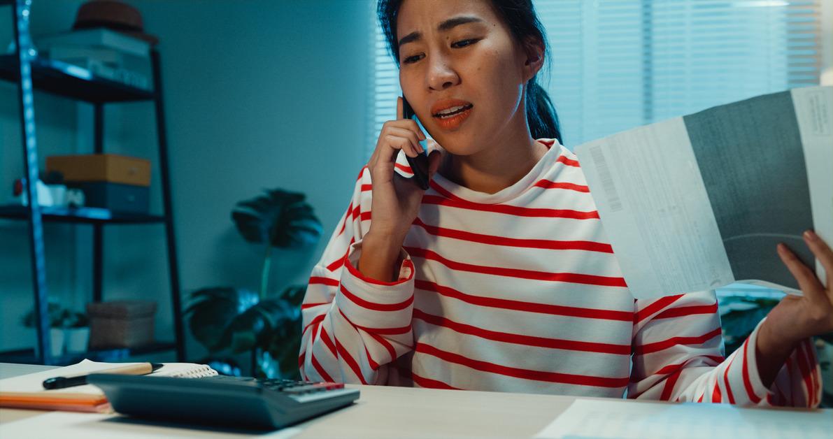 Woman in a red and white striped shirt looks frustrated while on the phone and holds a piece of paper. 