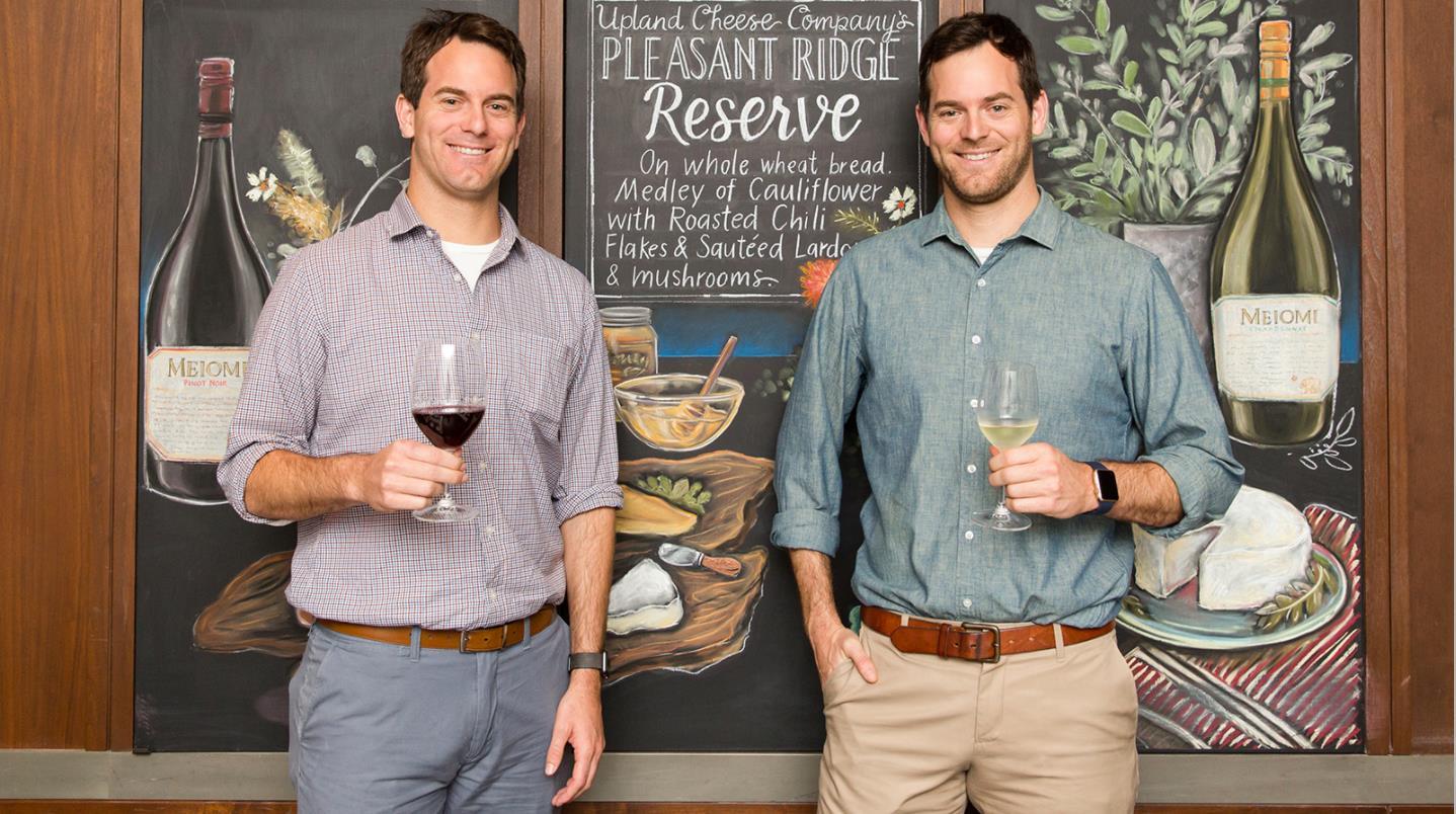 Michael and Charlie Kalish, known as the "Cheese Twins" on social media, are seen here at a wine tasting while wearing violet and green shirts and off-gray and khaki slacks.