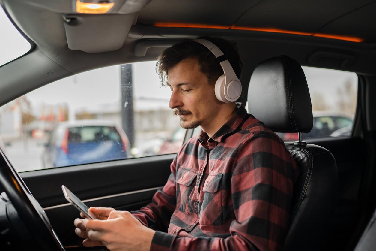 A man wears white headphones while using both hands to text while in the driver-side seat of a car.