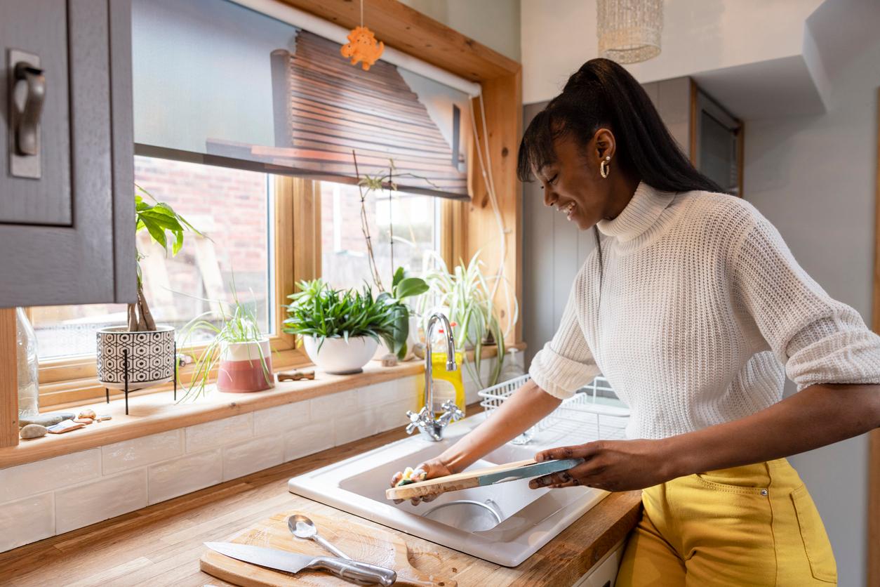 A smiling woman pre-washes her wooden cutting board without soap and water in her kitchen sink.