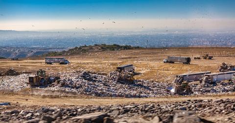 Trucks surround a landfill on the top of a sandy mountain while birds fly in the air.