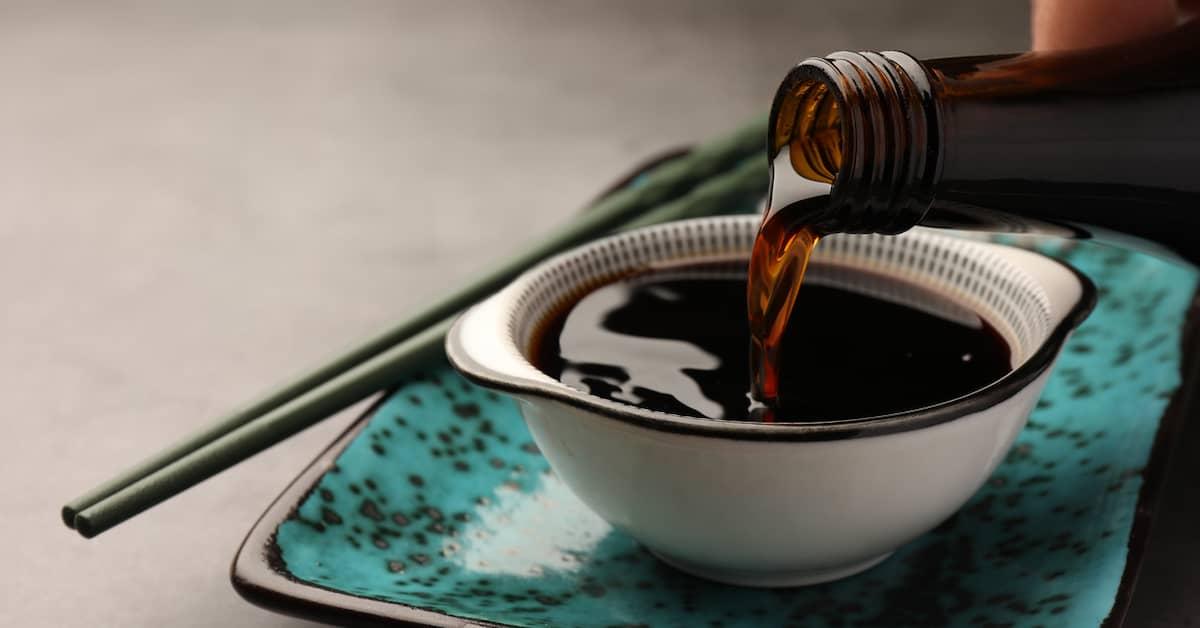 Soy sauce being poured into a white bowl standing on a blue plate with chopsticks.