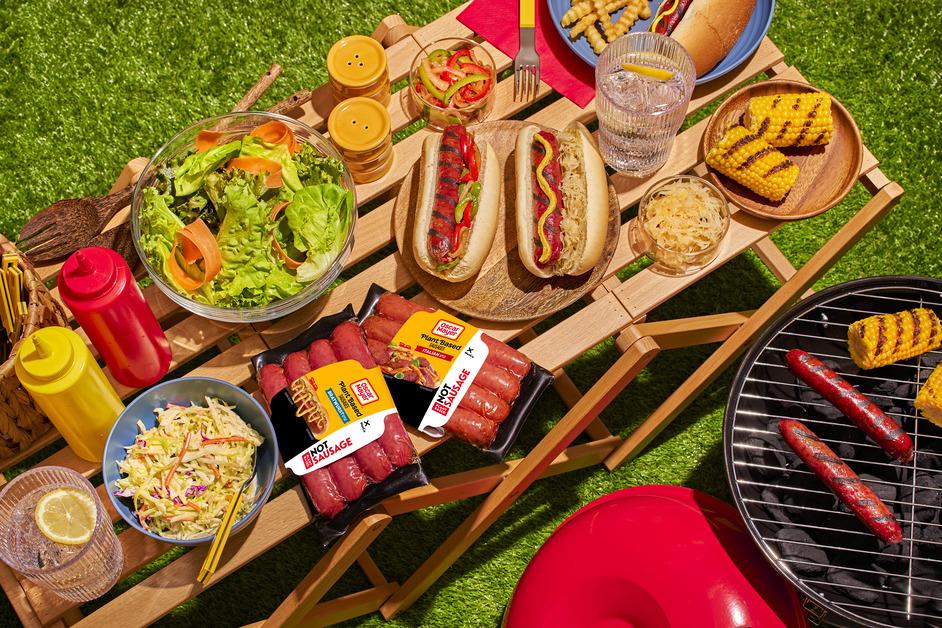 An overhead view of a picnic table covered in Oscar Mayer hot dogs, sausages, and other summer cookout foods next to a grill with corn and hot dogs grilling. 