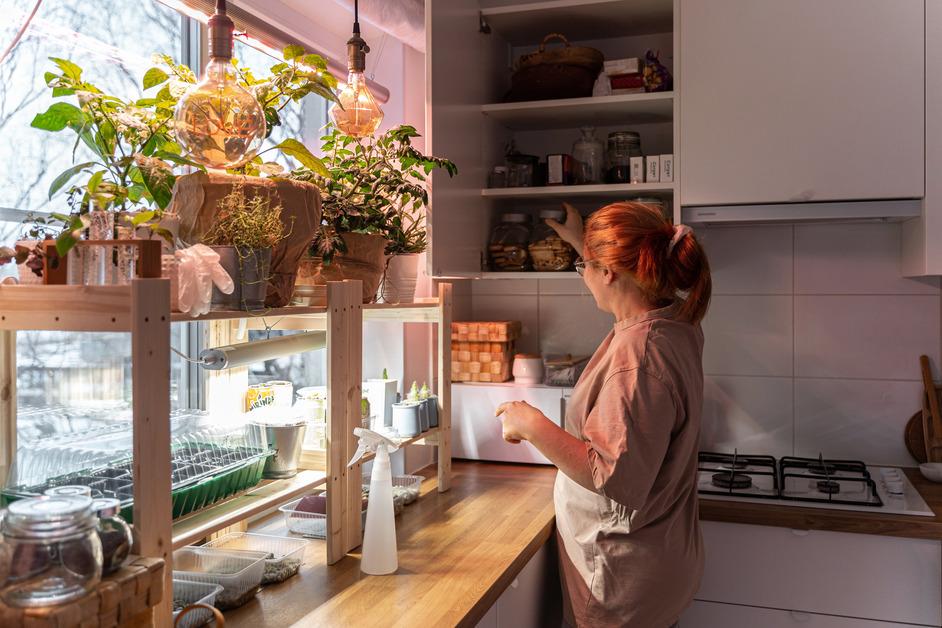 Woman reaches for something out of a cabinet in her kitchen next to a shelf of potted plants. 