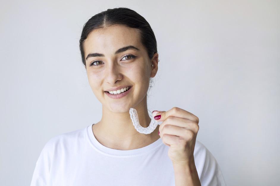 A woman wearing a white shirt holds her nightguard to her mouth and smiles.