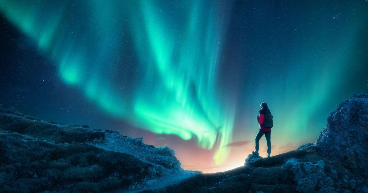 Woman standing on a rugged cliff and beholding a dazzling display of glassy blue-green auroral lights dancing in the night sky (Representative Cover Image Source: Getty Images | Den Belltsky)