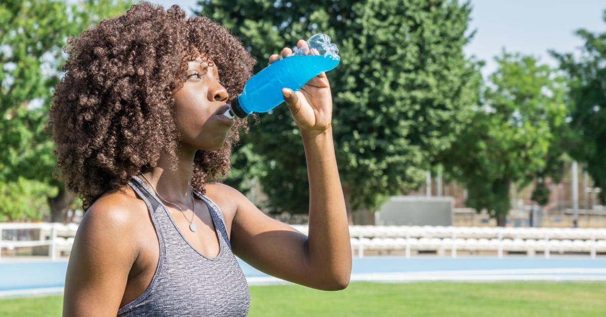 A woman wearing a gray tank top drinks a blue sports drink outside. 