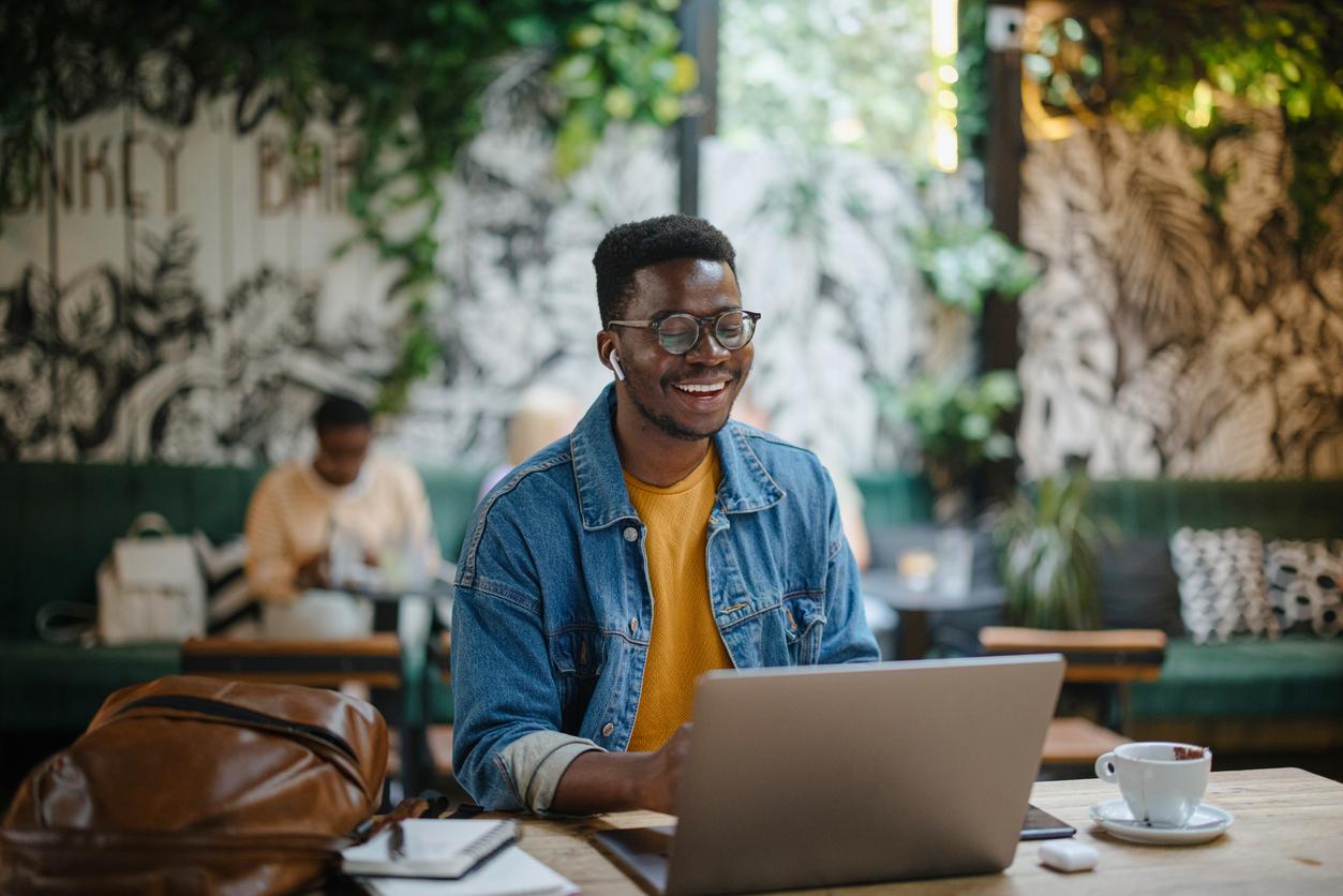 A smiling man looks down at his laptop while working inside a cafe.