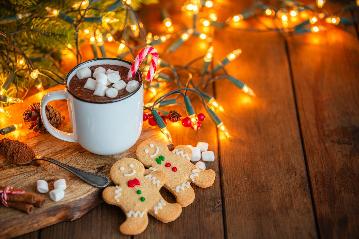 Two gingerbread cookies appear next to a cup of hot chocolate and holiday lights above a wood floor.