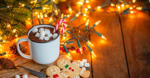 Two gingerbread cookies appear next to a cup of hot chocolate and holiday lights above a wood floor.