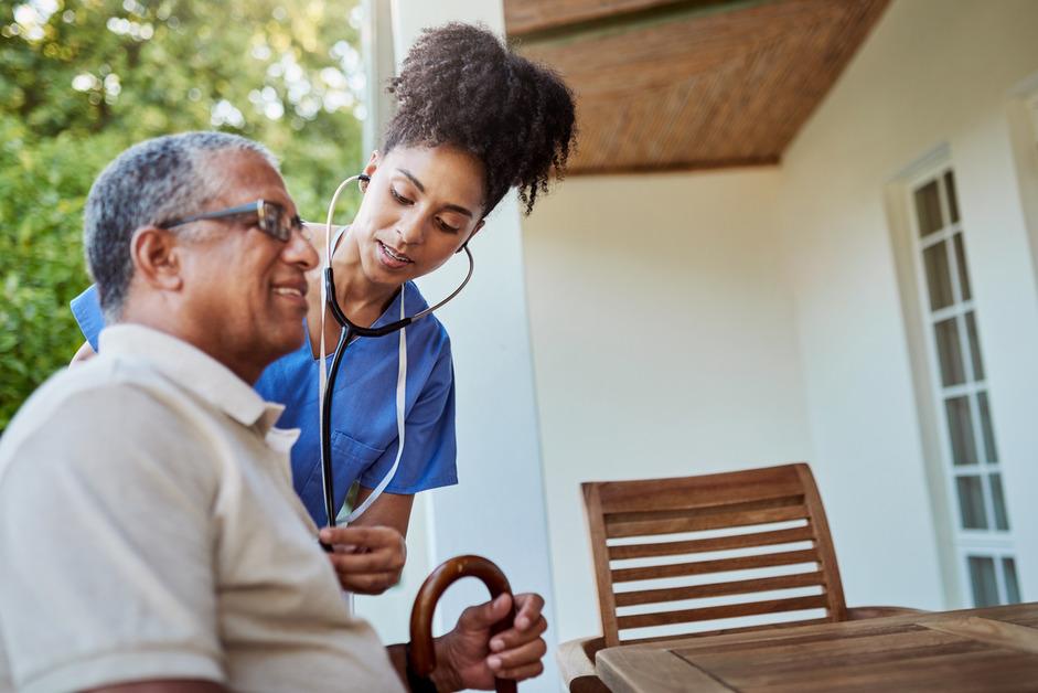 A man sits outside at a table while a nurse with a stethoscope stands next to him to check his heartbeat. 