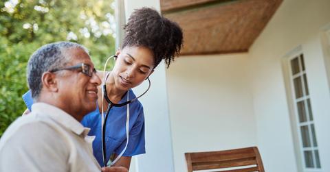A man sits outside at a table while a nurse with a stethoscope stands next to him to check his heartbeat.