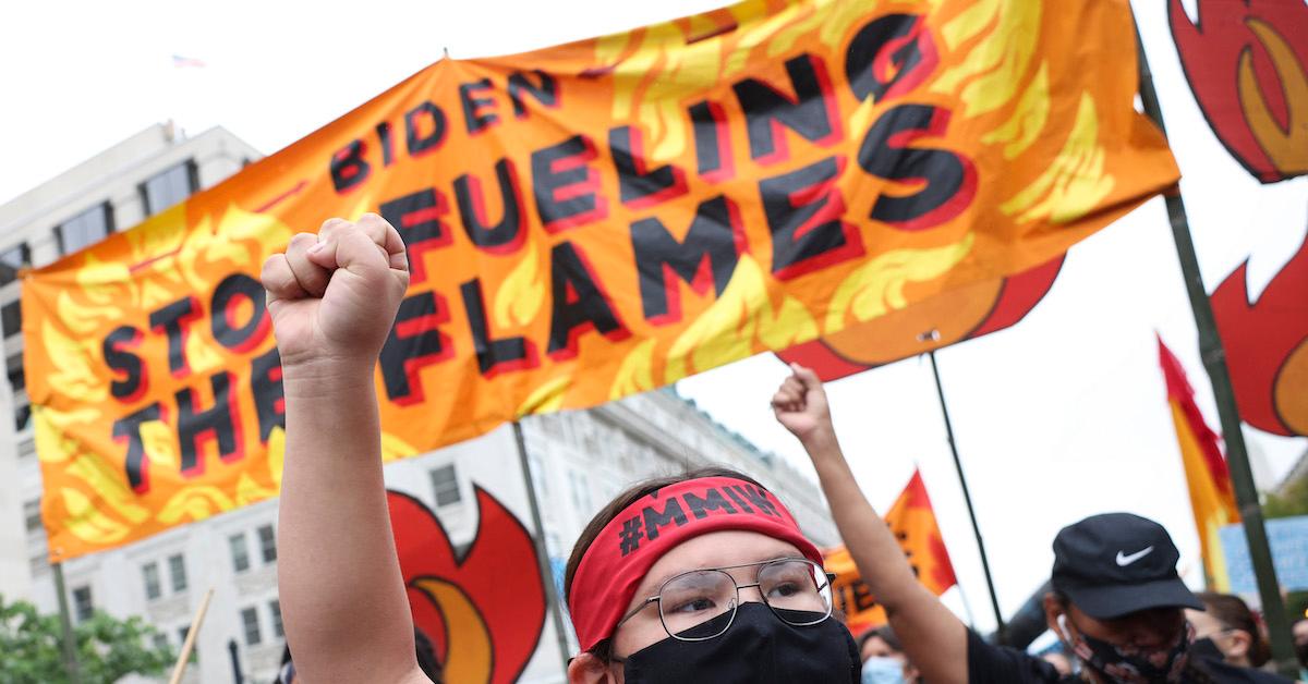 A protester wearing a "#MMIW" bandana holds up a fist, in front of an orange banner that reads "Biden: Stop fueling the flames."