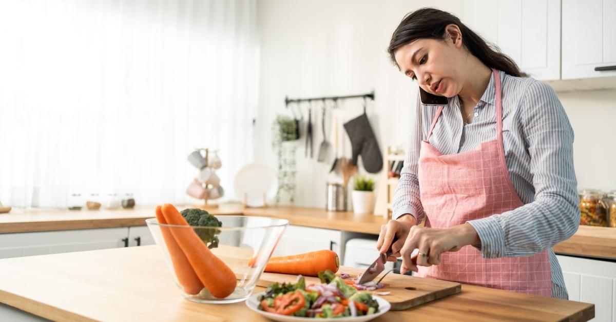 A woman talks on the phone while cutting large carrots into a salad in her kitchen. 