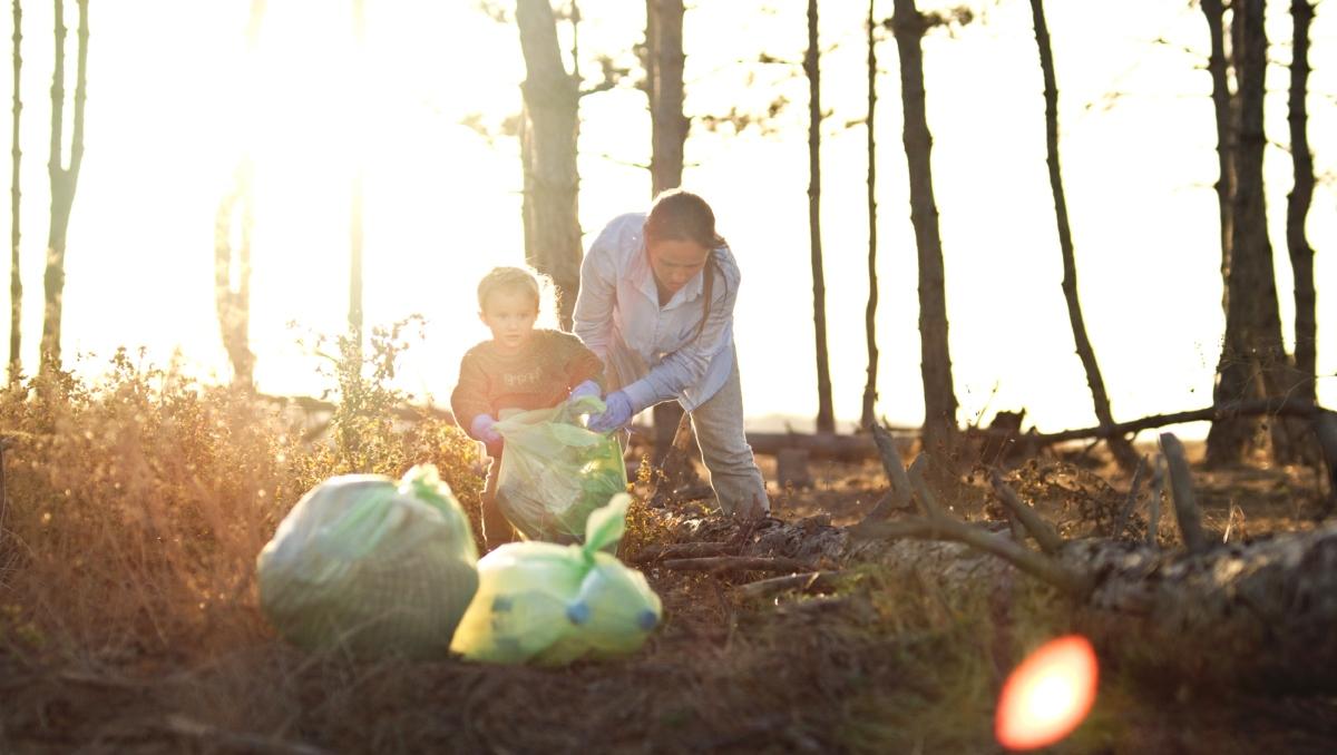 A little boy and woman pick up trash in the woods with the sun setting in the background. 