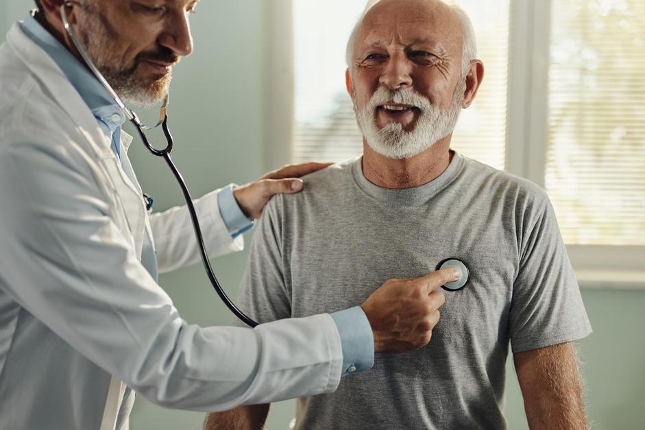 A senior man wearing a gray shirt sits while a cardiologist uses a stethoscope to check his heart. 