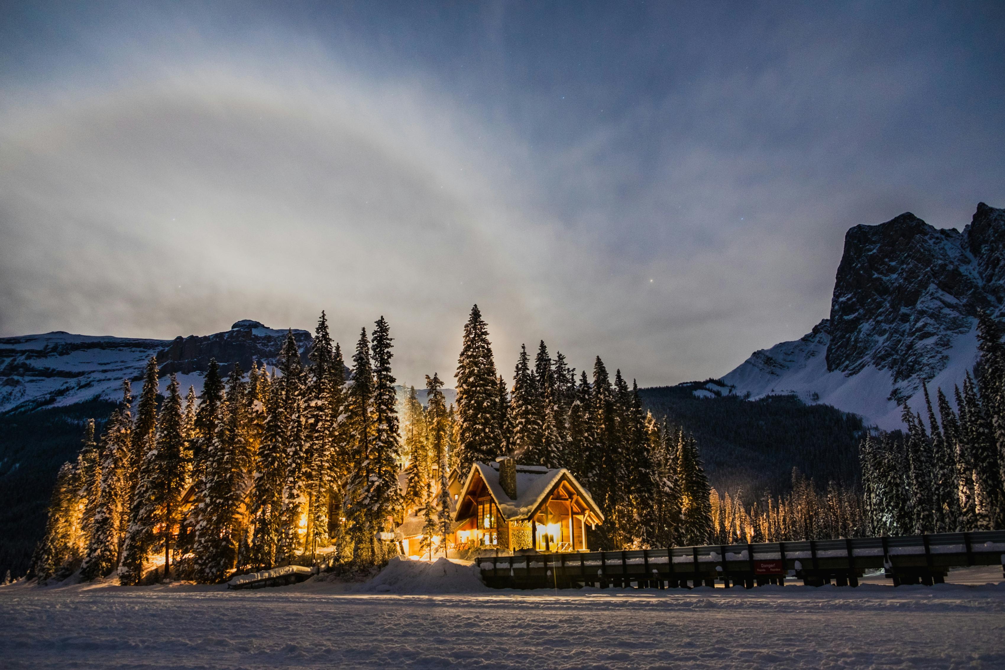 A house and surrounding trees are illuminated beside snowy mountains atop a snowy ground.