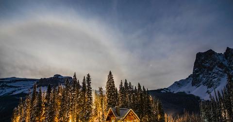 A house and surrounding trees are illuminated beside snowy mountains atop a snowy ground.